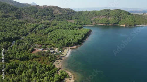 Aerial drone view of a small campsite and RV parking area on the forested shores of Lake Komani, Albania, featuring turquoise water and rugged mountain peaks in the Balkan wilderness.