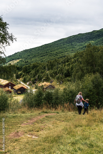 A mother and her child walk along a hiking trail in Innerdalen valley. Traditional log cabins with sod roofs are visible on the hillside in the background.
