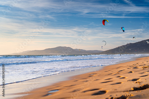 Fotografie Kites fly against the sky at sunset on coast with mountains in the background at famous surfing spot in Europe, Spain, Tarifa