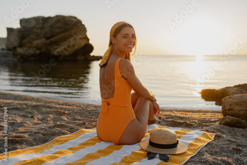 beautiiful smiling happy young woman sitting on beach by sea tanning dressed in orange swimsuit, summer fashion vacation accessories trend