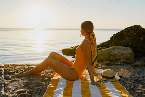 beautiiful smiling happy young woman sitting on beach by sea tanning dressed in orange swimsuit