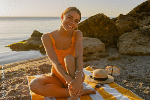 beautiiful smiling happy young woman sitting on beach by sea tanning dressed in orange swimsuit