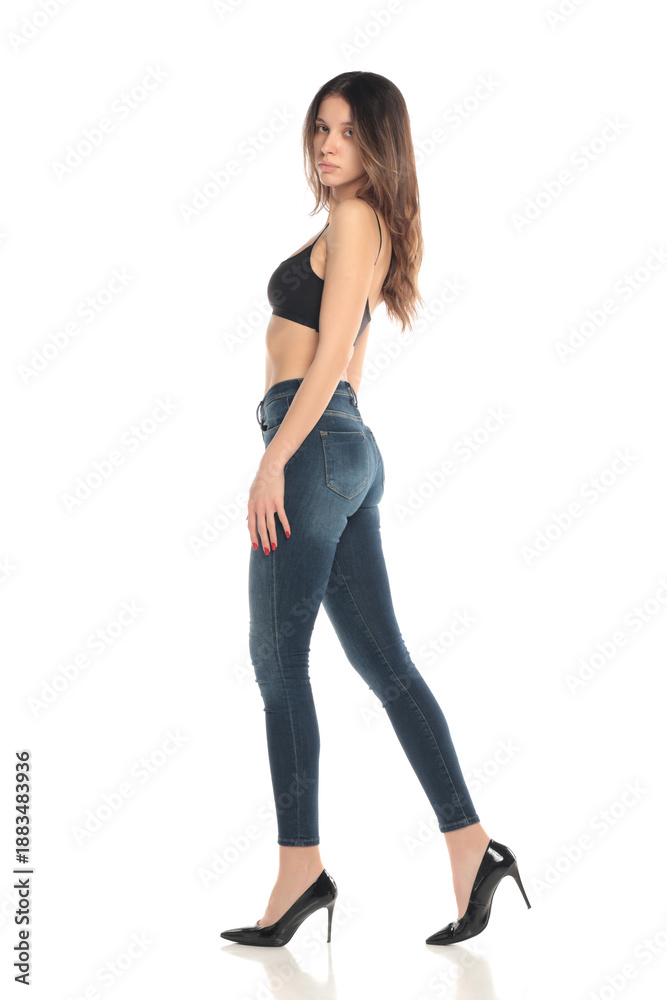 Fototapeta premium Side view full body studio shot of a young woman in black bra and jeans standing in profile on a white background