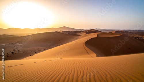 Vast dunes roll under the warm glow of a sunrise, casting shadows and revealing the textured landscape. Mountain silhouettes