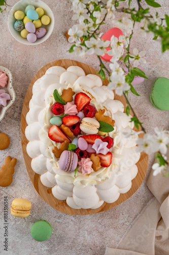 Easter egg-shaped Pavlova cake with whipped cream, topped with fresh berries and assorted sweets on a wooden board on a concrete background. Top view, copy space.