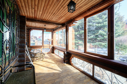 Fotografi Wooden veranda with large panoramic windows, log ceiling, wicker chair, decorative wrought iron railings, lantern lighting — bright, airy, cozy space with garden views