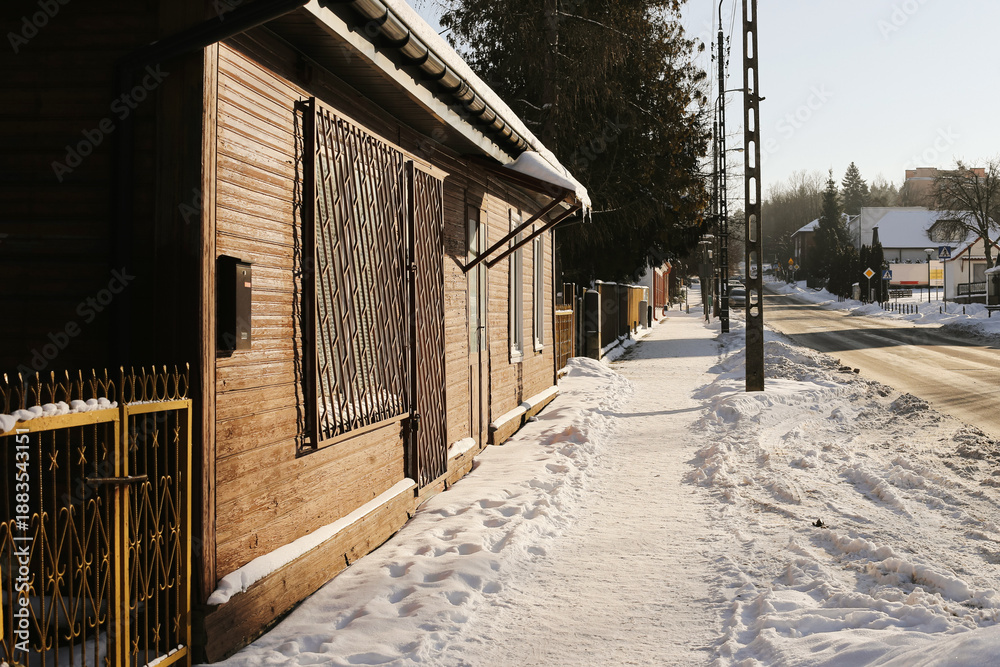 Naklejka premium Snowy residential street with sidewalk and houses. Winter neighborhood scenery with icy pavement and sunlight. Quiet suburban area during cold weather. Urban daily life in winter season.