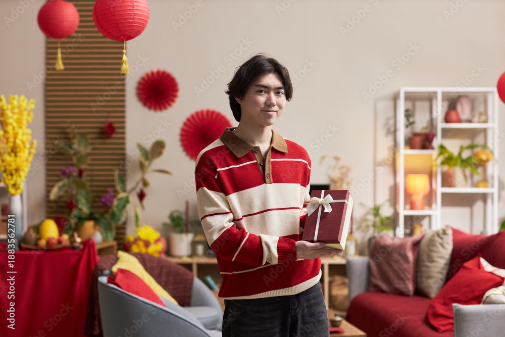 Fototapeta premium Portrait of young adult Asian man smiling and holding gift box, while standing in decorated living room with Tet holiday ornaments and festive plants visible in background