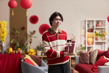Portrait of young adult Asian man smiling and holding gift box, while standing in decorated living room with Tet holiday ornaments and festive plants visible in background