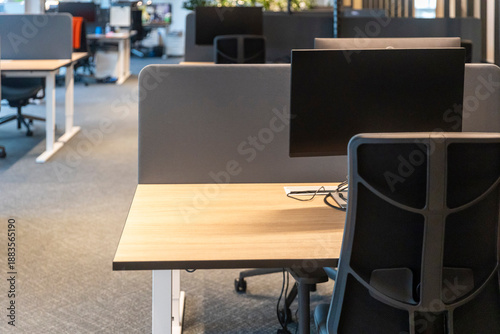 Contemporary office environment featuring wooden desks with monitors, ergonomic black office chairs, white privacy screens, and modern minimalist interior design with indirect lighting.