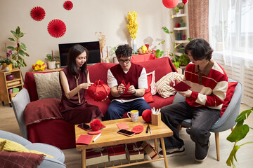 Three Asian young adults sitting on sofa preparing red Tet holiday decorations, holding paper crafts and yarn, engaging in traditional festive activity in living room setting