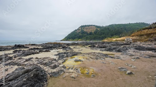 A wide shot of the dramatic and rugged coastline at Cape Perpetua Scenic Area near Yachats, Oregon. The scene features dark volcanic rock formations, tide pools, and a sandy beach foreground, with the