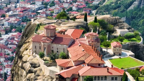 Cinematic aerial orbit of St. Stephen's Monastery in Meteora, Greece. Drone footage reveals the stunning Orthodox complex, gardens, and red roofs against the dramatic cliffs