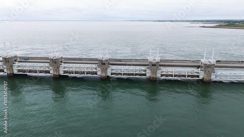 massive white steel truss gates storm surge barrier integrated into thick concrete supports these structures span across dark green coastal waters under hazy sky netherlands delta works 