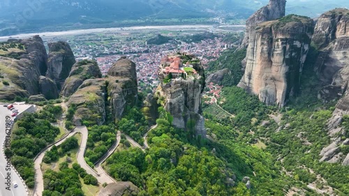 Wide aerial drone shot approaching the Holy Trinity Monastery in Meteora, Greece. Establishing shot revealing the winding roads, cliffs, and Kalabaka town in the valley below