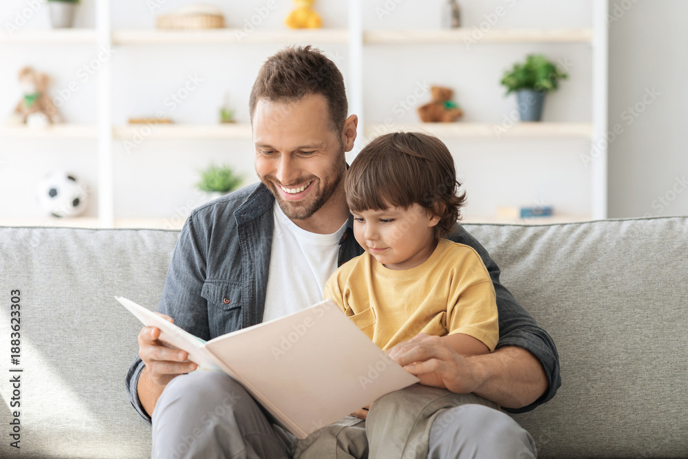 Fototapeta premium Weekend activities with toddlers. Cheerful carying daddy reading book to little son, baby boy enjoying fairy tale on dad's knees, sitting on sofa at home