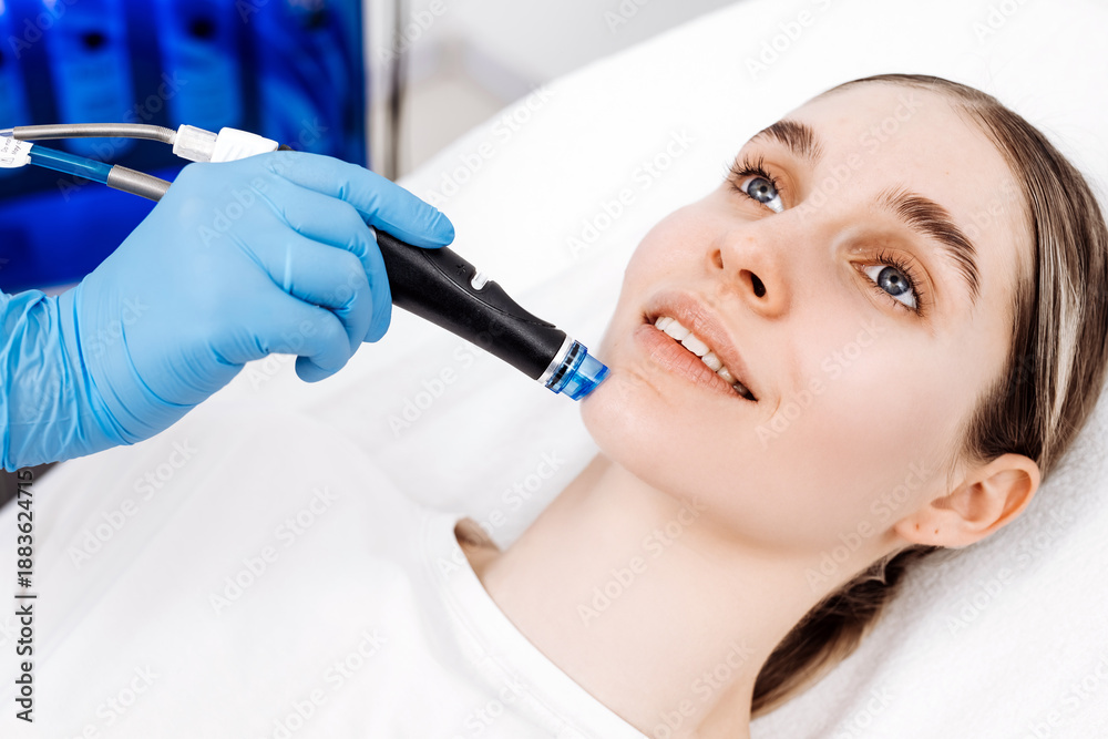 Naklejka premium Cosmetologist performs a hydrafacial treatment on a smiling client in a skincare clinic. Concept skincare, facial treatment, beauty care