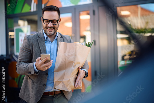 Fotografia Happy man carrying paper bag full of groceries while using cell phone in front of supermarket