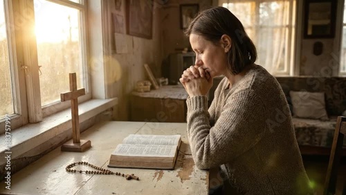 Christian woman praying with rosary and open bible at rustic table next to window during sunset