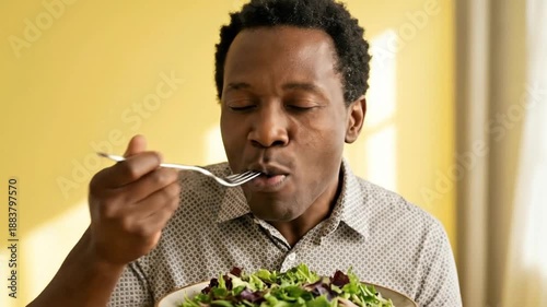 Happy African American man eating fresh green salad and savoring healthy vegetarian meal on yellow background