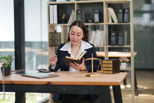 Asian businesswoman or lawyer in a formal blue suit sitting at a wooden desk, using a digital tablet. The desk is decorated with legal symbols including a gold scale of justice and law books.