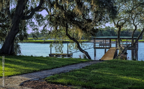 Sunny morning view of a path leading to a dock house and pier built on a grassy shoreline, with several trees as well as marshes visible