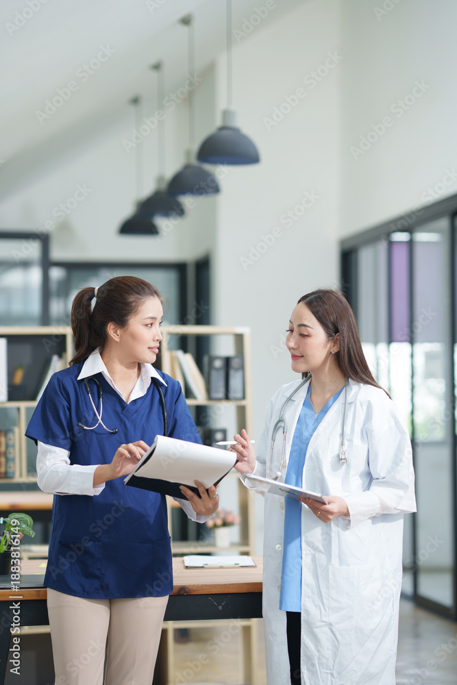 Obraz premium female doctor in a white lab coat and a medical assistant in blue scrubs are sitting at a wooden desk, collaborating and reviewing medical documents or X-rays. Both are wearing stethoscopes, reflectin