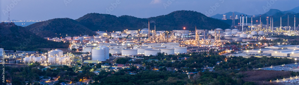 Naklejka premium Panoramic night view of a large oil refinery and petrochemical plant located near mountains. Industrial energy estate landscape with illuminated storage tanks and distillation towers at twilight.