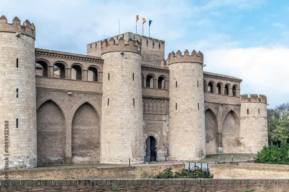 Fototapeta premium Historical facade of Aljaferia Palace showing intricate Mudejar arches and round stone towers in Zaragoza. A prime example of Islamic architecture in Spain under a soft daylight sky