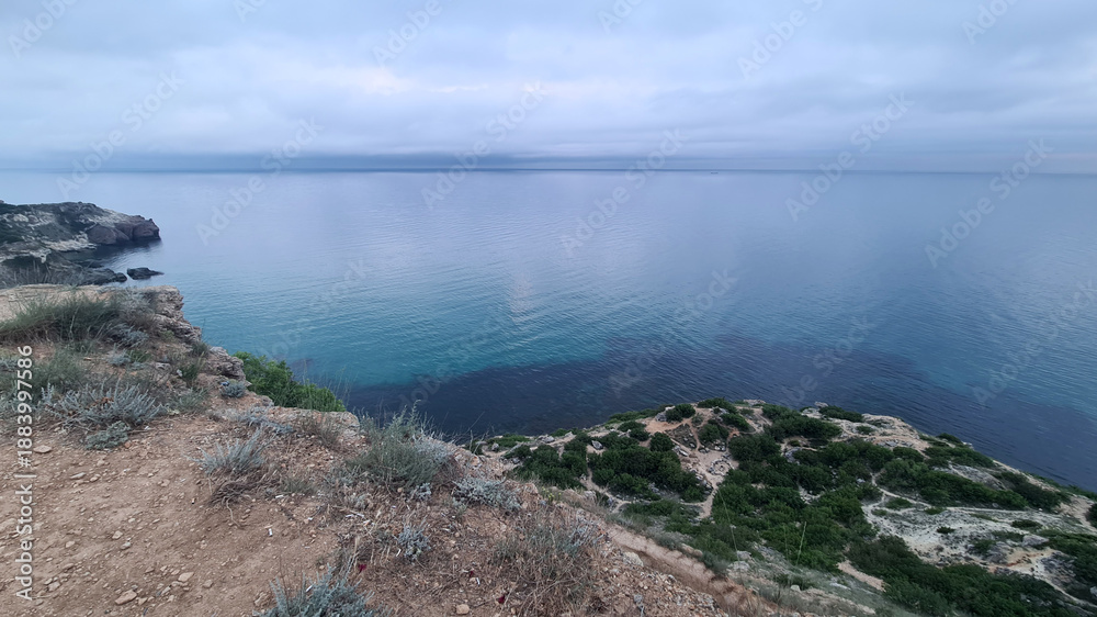 Fototapeta premium A serene coastal landscape featuring a rocky cliff overlooking calm blue waters. The sky is overcast, creating a tranquil atmosphere.