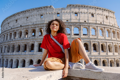 A young woman beaming with joy sits on a stone ledge, with the majestic and iconic Colosseum in Rome proudly displayed behind her, perfectly showcasing the charm and allure of this historic city