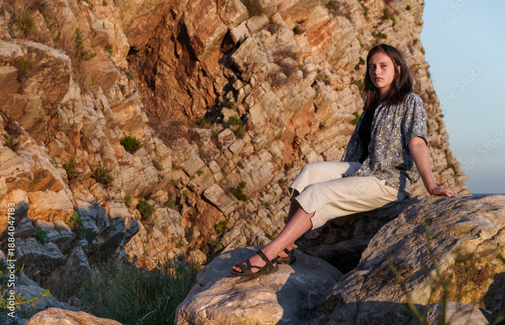 Naklejka premium A girl poses by the sea, standing on rocks near the slope of a high cliff, panoramic view of the seashore and mountains at sunset
