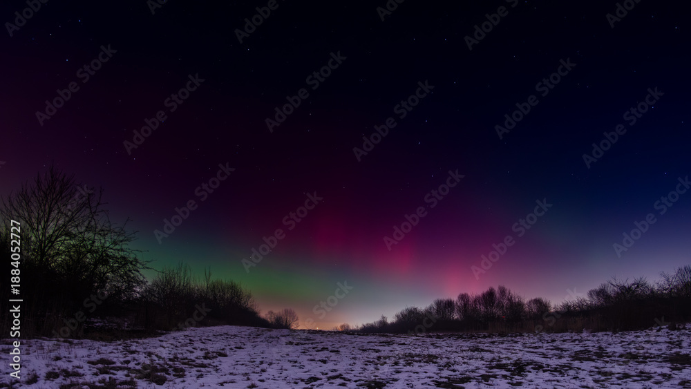Fototapeta premium Vivid aurora borealis illuminating the winter night sky above snow-covered open field in Silesia region of Poland, with intense green and magenta light pillars, 19.01.2026