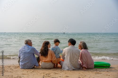 Happy multi-generational Asian family sitting on the beach with their backs to the camera, raising hands and enjoying fun vacation moment together by the sea. family, beach, vacation, summer. elderly