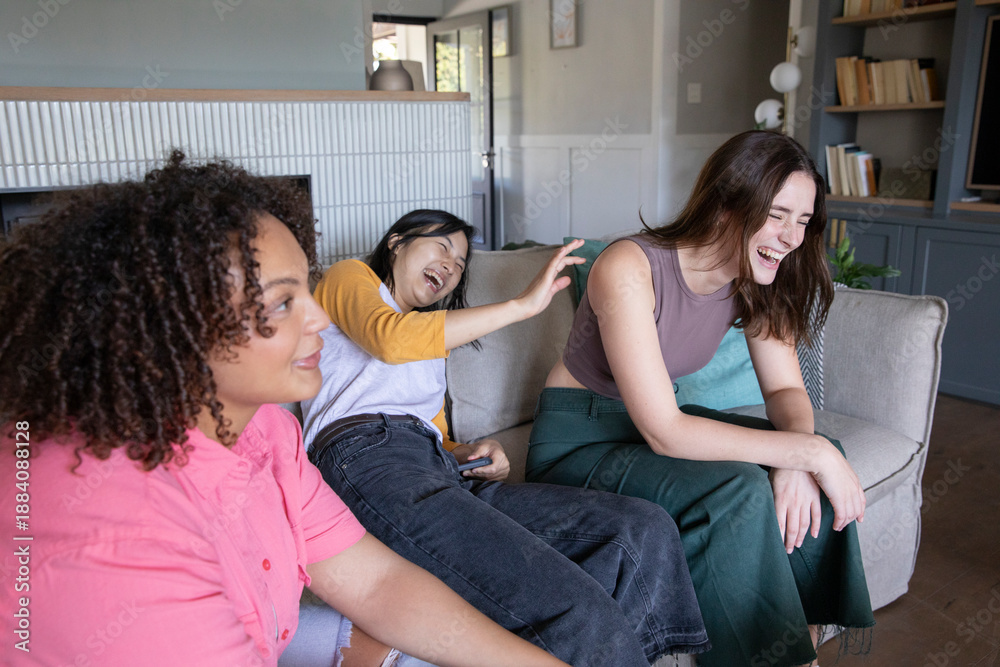 Fototapeta premium Diverse female friends laughing together on upholstered couch in living room holding remote control