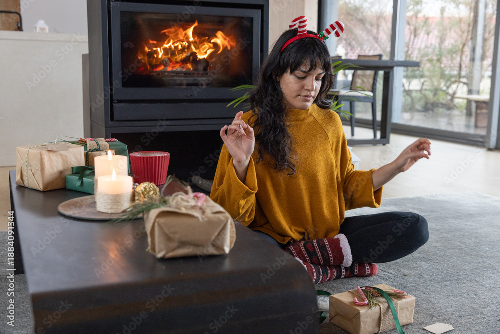 Obraz premium Woman sitting cross-legged on gray rug next to fireplace with gifts and candles, copy space