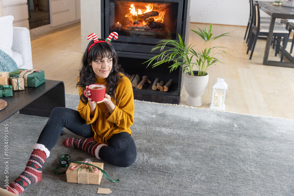 Fototapeta premium Woman wrapping christmas gifts on gray rug in living room next to burning fireplace with red mug