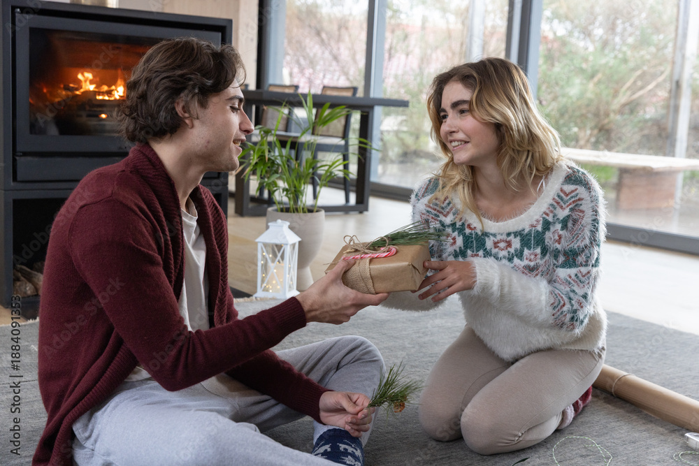 Naklejka premium Couple kneeling and exchanging craft-wrapped gift with pine sprig on grey rug by lit fireplace