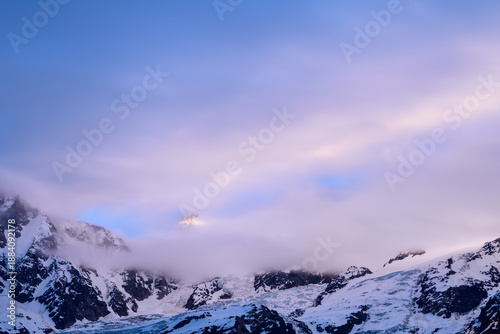 Fotografie A snow-covered alpine peak briefly catches golden sunlight as soft pastel clouds drift across the Mont Blanc du Tacul massif, creating a serene and ethereal mountain scene