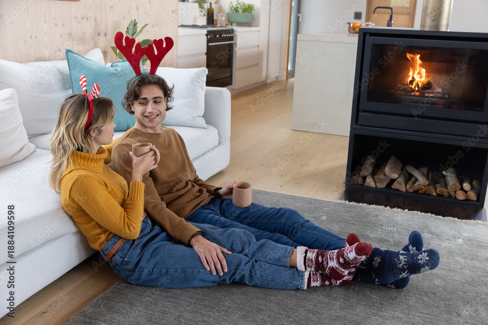 Obraz premium Couple sitting on gray rug in living room beside fireplace holding mugs wearing antler headbands
