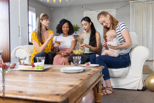 Foto Diverse female friends sitting in living room holding infant, eating snacks arou