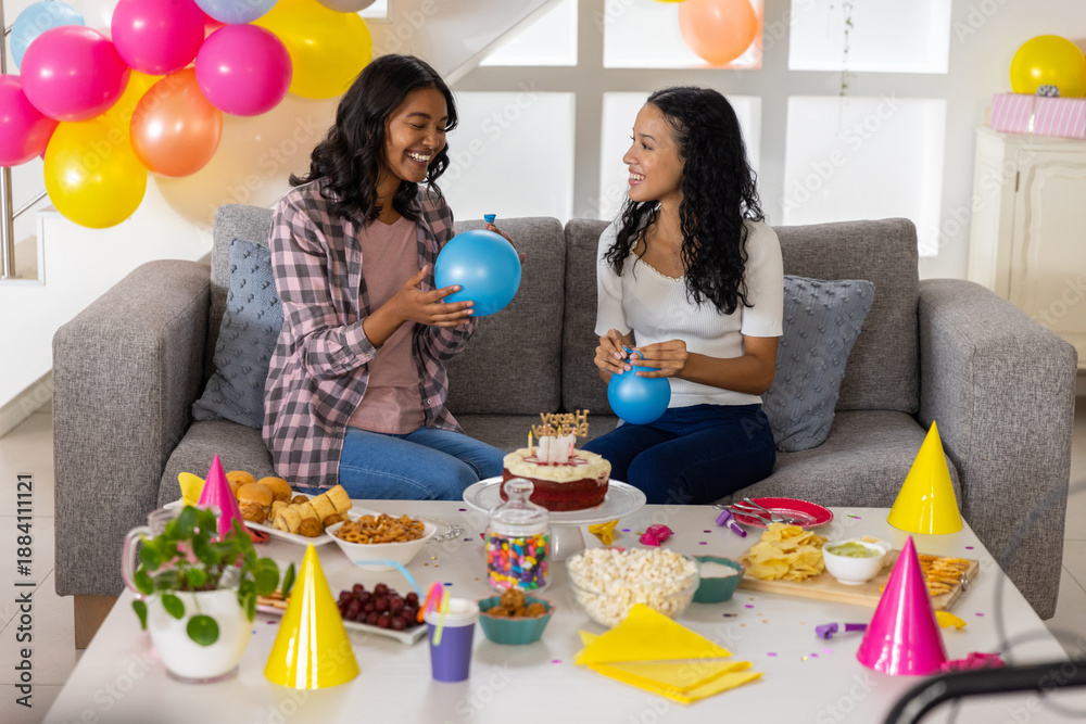 Obraz premium Diverse mom and teenage daughter sitting on sofa holding blue balloons amid birthday cake, popcorn