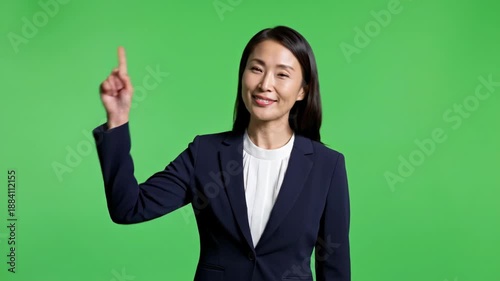 An Asian woman in a dark suit points up with a smile on a green backdrop