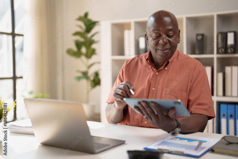 Fototapeta premium Mature African American businessman analyzing data on a digital tablet while working remotely from a modern home office, symbolizing modern work, technology, and success