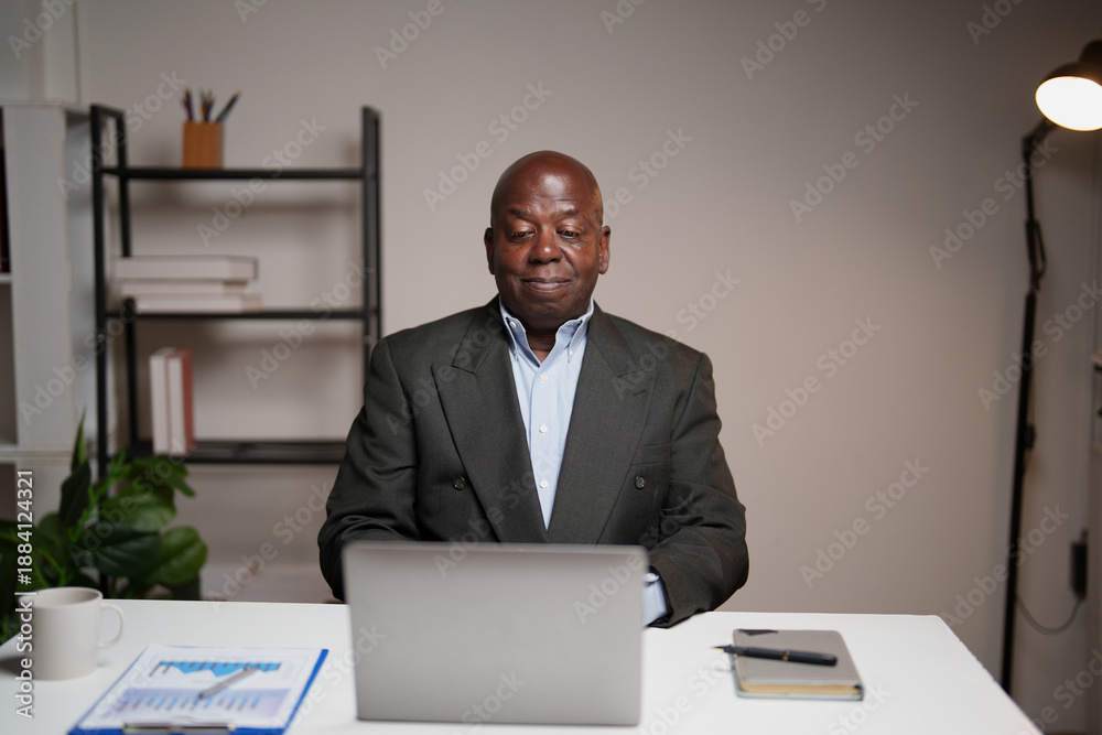 Naklejka premium Senior African American executive sitting at a desk and using a laptop, performing professional tasks and managing business operations in a contemporary workspace