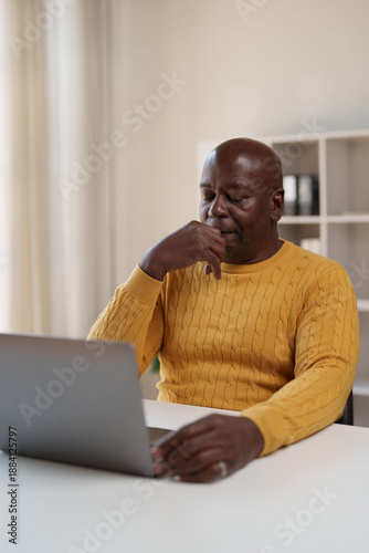 Mature Black man working from his home office, pensive and stressed, resting chin on hand as he concentrates on a laptop screen while tackling a difficult work problem