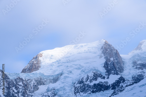 Soft, diffused sunlight gently illuminates the snow-capped summit and rugged ice formations of Mont Blanc du Tacul, with a tranquil blue sky and subtle mist creating a serene alpine atmosphere.
