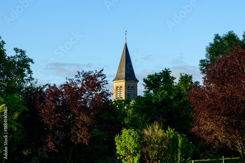 Фотография A tall church steeple emerges from dense green and burgundy trees under a clear blue sky in Saint-Honore-les-Bains, capturing a peaceful rural atmosphere