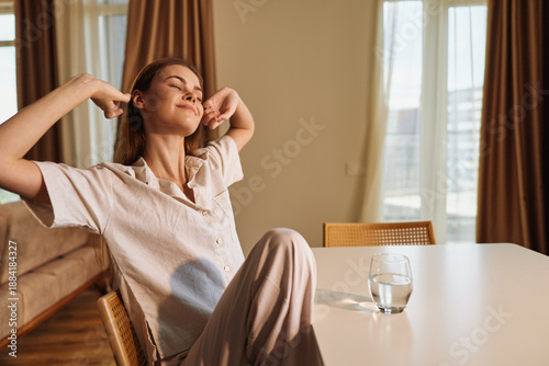 фотографія Woman morning stretching and relaxation at home in sunlight, sitting at table with glass of water, casual loungewear and peaceful wellbeing, enjoying quiet slow routine