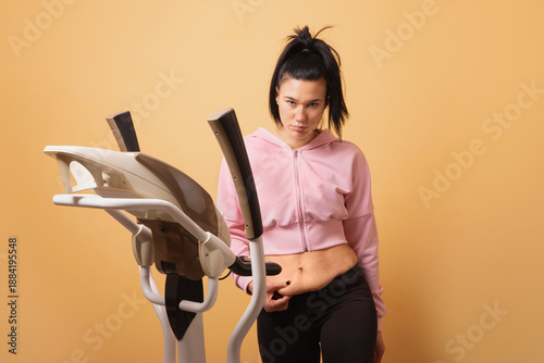Obraz na plátně A young woman in sportswear stands by an elliptical trainer and gently shows loose stomach skin against a solid yellow background
